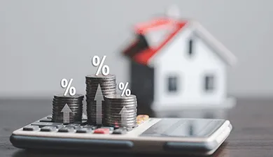 A house in the background and a calculator with coins stacked atop it